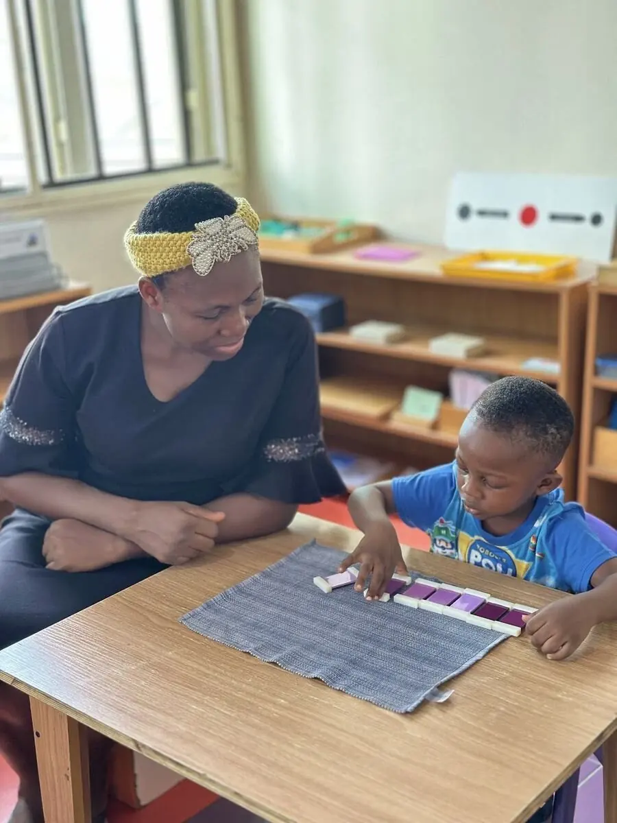 An AMI-certified guide sitting beside a child working with Montessori materials